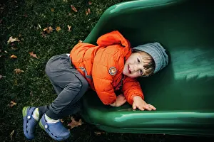 Whimsical Prints: Smiling child in orange coat lying on a green playground slide
