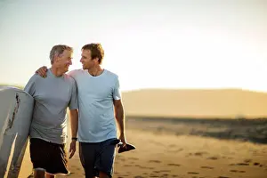 30 To 35 Years Prints: Smiling father and middle aged son walking along a beach with their paddleboard
