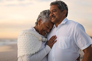 Biracial Prints: Smiling senior biracial woman embracing man looking away while standing at beach during sunset