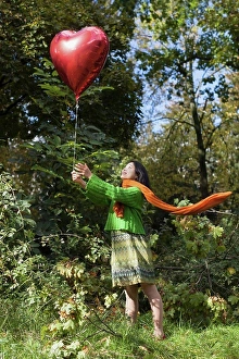 Sun Light Prints: Smiling woman holding red heart shape balloon while standing on field against trees at park