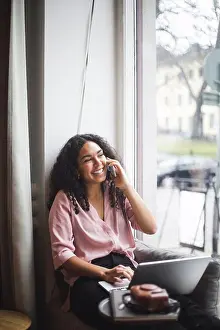 Communications Prints: Smiling young female entrepreneur talking through smart phone sitting on window sill in office