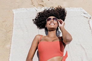 Grownups Prints: Smiling young woman with curly hair wearing sunglasses while lying on beach towel