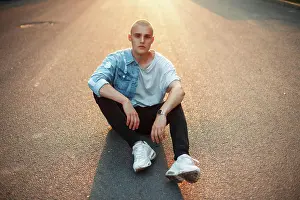 Stadt Prints: Stilvoller junger brutaler Mann mit Glatze in trendiger Jeanskleidung sitzt bei Sonnenuntergang auf dem Asphalt ***