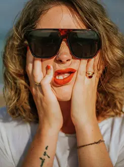 Individuality Prints: Stylish brown curly haired woman with red lipstick in trendy sunglasses looking at camera while squeezing face