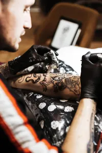 Focused Prints: From above stylish man with piercing using tattoo machine to make tattoo on leg of crop customer during work in salon