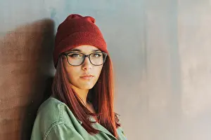 Pensive Prints: Thoughtful stylish woman in warm hat and glasses in dark green shirt nearby brown wall looking along