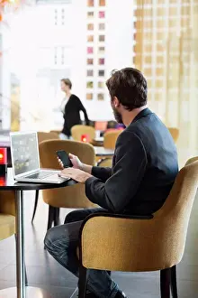Communications Prints: Side view of businessman with mobile phone and laptop sitting in hotel restaurant