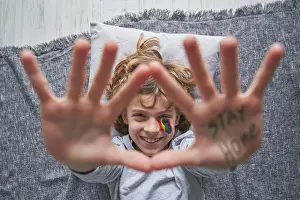 Inscription Prints: Top view of happy boy with colorful rainbow under eye showing stop gesture with hands with stay home inscription to