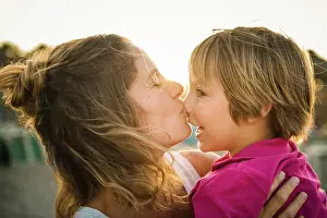 Carry Prints: Side view of laughing woman carrying cheerful playful son on hands while kissing his nose on beach in sunset