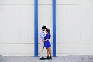 Architectural Prints: Side view of loving man and woman in elegant clothes kissing on background of white wall with bright blue columns