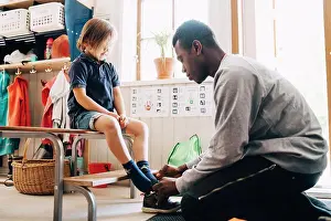Midadult Men Prints: Side view of male teacher helping boy in wearing shoe at kindergarten