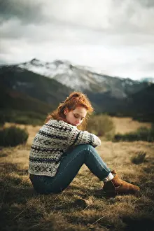 Sweater Prints: Side view of pensive charming woman with red hair in warm sweater sitting with closed eyes on lawn in mountain in gray
