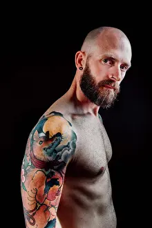 Macho Prints: Side view of shirtless bald serious hipster with tattoos on hands looking at camera on black background