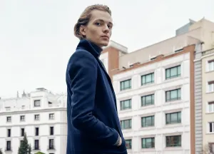 Apparel Prints: Side view of stylish young male with trendy hairstyle dressed in elegant coat standing against gray stone wall in city