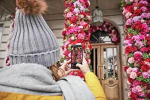 Anonymous Prints: Back view of unrecognizable female in outerwear using smartphone to take photo of beautiful floral decorations outside