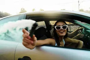 Road Trip Prints: Side view of young cheerful girl in sunglasses riding in car and holding gas bomb