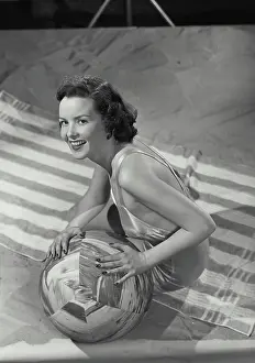 Suit Prints: Vintage photograph. Brunette woman smiling wearing bathing suit sitting on beach towel with ball