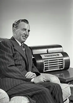 Unwind Prints: Vintage Photograph. Caucasian man wearing pin striped suit sitting in arm chair next to large radio