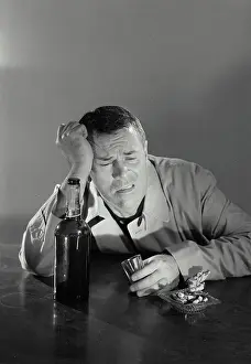 Cigarettes Prints: Vintage Photograph. Drunk man leaning on elbow on table with liquor bottle and shot glass and ashtray with cigarettes