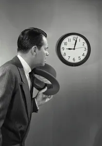 Deadline Prints: Vintage photograph. Man with dark hair and suit holding hat standing next to wall clock