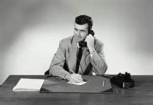 Notes Prints: Vintage Photograph. Man sits at desk taking notes on small card smiling he talks on phone
