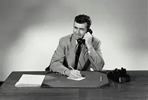 Notes Prints: Vintage Photograph. Man sits at desk taking notes he smiles with a concerned look on his face he talks on phone