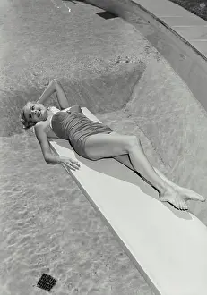 Suit Prints: Vintage Photograph. Smiling blonde woman wearing bathing suit lying on diving board over swimming pool