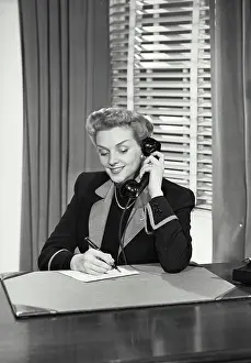 Desk Prints: Vintage Photograph. Woman at desk talking on telephone and writing on paper