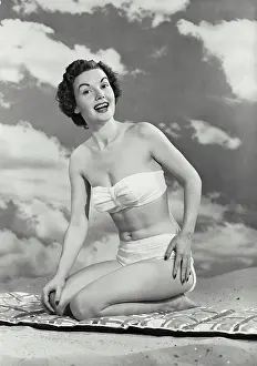 Bikini Prints: Vintage photograph. Woman smiling in swimsuit kneeling on mat in sand