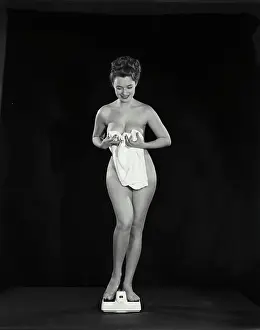 Vintage Photograph. Young nude woman with bath towel standing on scale in front of black background