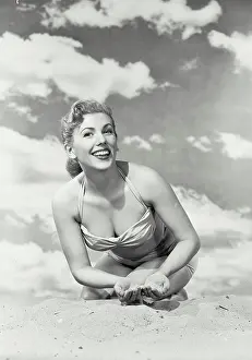 Bikini Prints: Vintage photograph. Young smiling woman in swimsuit kneeling in sand with sky background