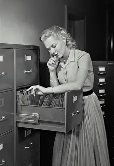 File Prints: Vintage Photograph. Young woman wearing dress going through folders in drawer of filing cabinet