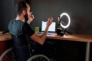 Disability Prints: Vlogger filming through mobile phone with circular strobe tripod while sitting on wheelchair at studio