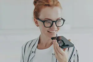 Conversation Prints: Voice search. Close up shot of redhead woman in transparent glasses speaks to virtual assistant holds mobile phone near