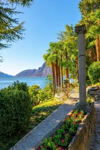 : Walkway with Column on Lake Lugano with Mountain Peak and Palm Tree in a Sunny Day with Clear Sky in Lugano