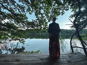 Kindness Prints: woman in skirt looks out from wooden platform over green natural lake