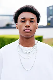 Apparel Prints: Young black curly haired male teenager in stylish clothes with chains looking at camera while standing on city street
