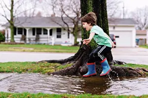Life Styles Prints: Young boy with brown hair jumping into a big puddle by the side of a street