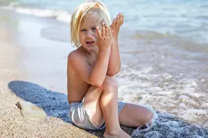 Unschuld Prints: A young child sitting on a sandy beach covering one side of their face with sand-covered hands