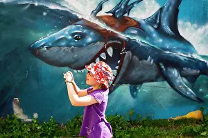 Humour Prints: Young girl holding camera standing in front of graffiti painting of shark that looks as if it is biting her head