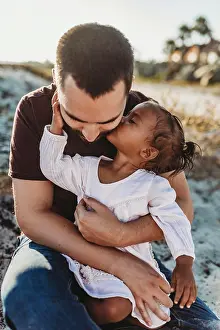 Person Of Color Prints: Young girl sitting in fathers lap giving a kiss at beach
