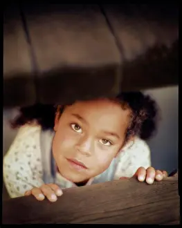 Verticals Prints: A young girl under a table sheepishly looks upward