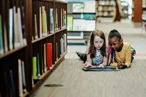Young Girls Prints: Two young girls reading a book on the floor of library