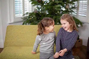 Sofa Prints: Two young girls sitting on sofa in living room, smiling at each other