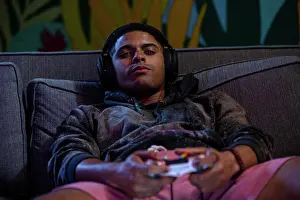 Arts Culture And Entertainment Prints: Young man laying back on sofa with video game controller in hand and headset