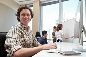 Place Of Work Prints: Young man using computer in office looking towards camera