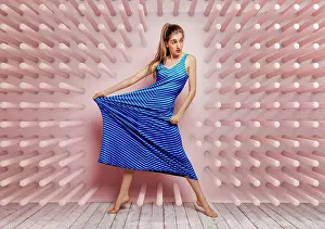Attractive Prints: Young woman barefoot looking away stretching side of a blue dress while standing on tube hook pink background