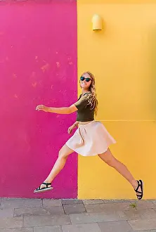 Overlooking Prints: Young woman in dress jumps happily in front of colorful house